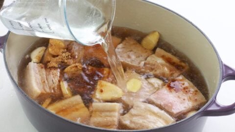 Adding water to a pot of pork belly cubes with garlic, ginger, and seasonings.