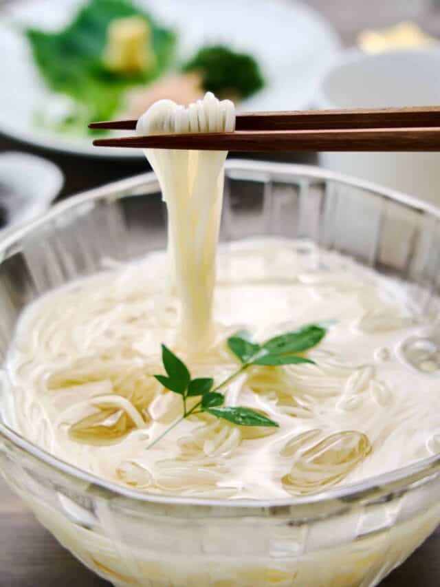 Close up of whisper thin strands of chilled somen noodles being lifted from a bowl of ice water set against a backdrop of vibrant condiments.