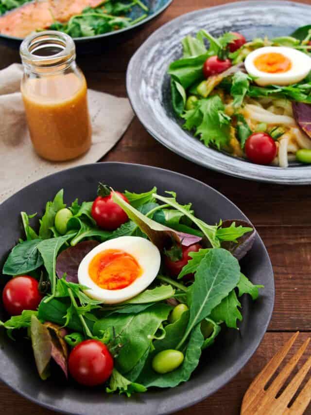 High-angle shot showing a vibrant table spread with a baby green salad, poached salmon, and cold udon salad, all enhanced by a bottle of Ginger Miso Dressing.