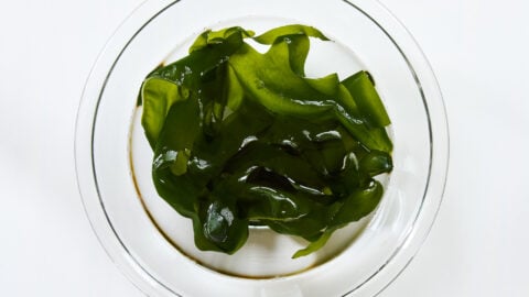 Glossy green ribbons of rehydrated wakame seaweed unfurls in a bowl.
