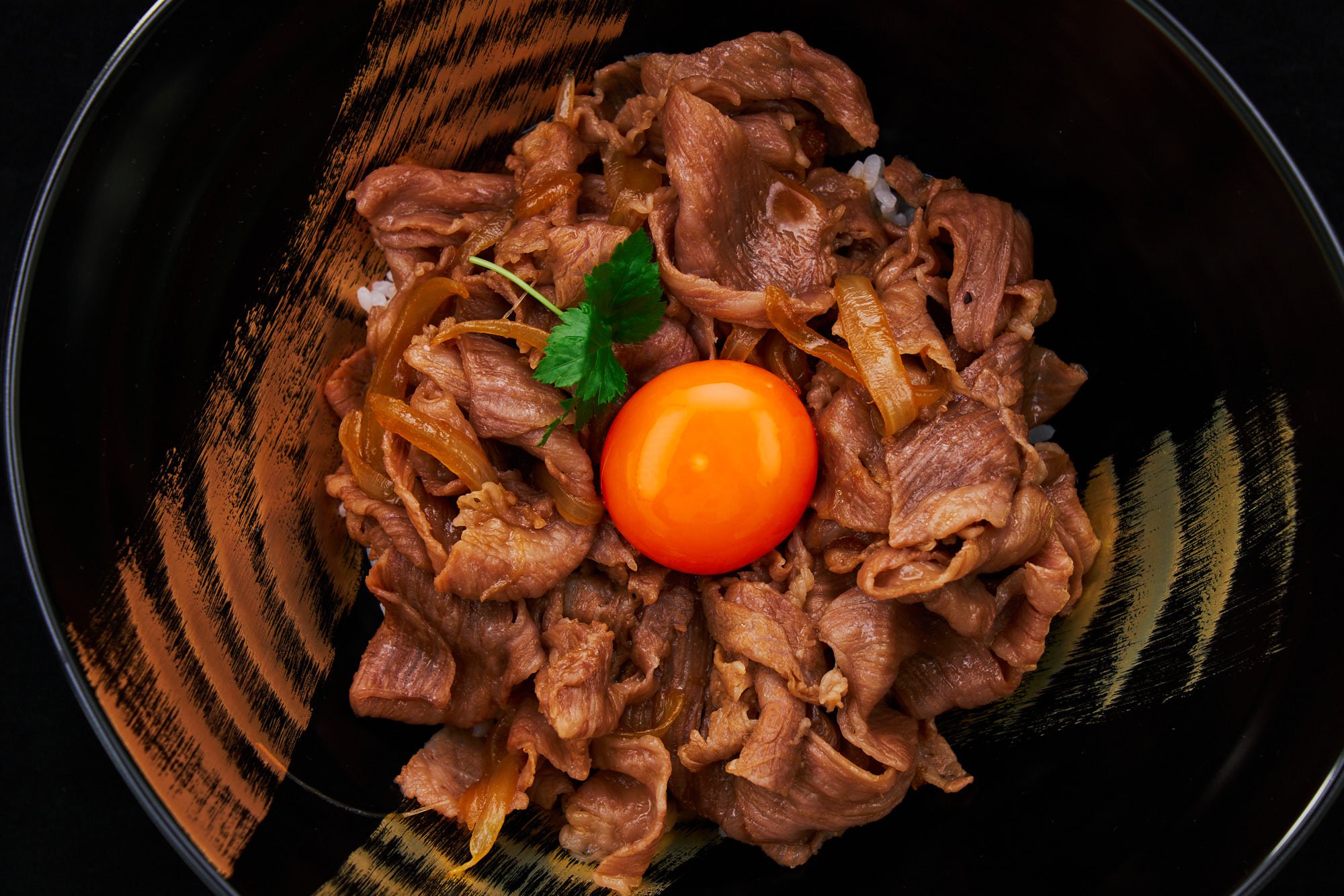 Overhead shot of a gyudon bowl reveals its balanced presentation. The beef's marbling shines through the glossy sauce, while the egg yolk provides a pop of color, perfectly framed by the luxurious black and gold bowl.