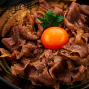 A stunning close-up of gyudon captures tender slices of perfectly cooked wagyu beef glistening with a rich, savory sauce. The vibrant orange egg yolk rests at the center.