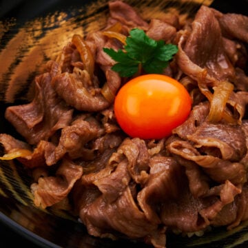 A stunning close-up of gyudon captures tender slices of perfectly cooked wagyu beef glistening with a rich, savory sauce. The vibrant orange egg yolk rests at the center.