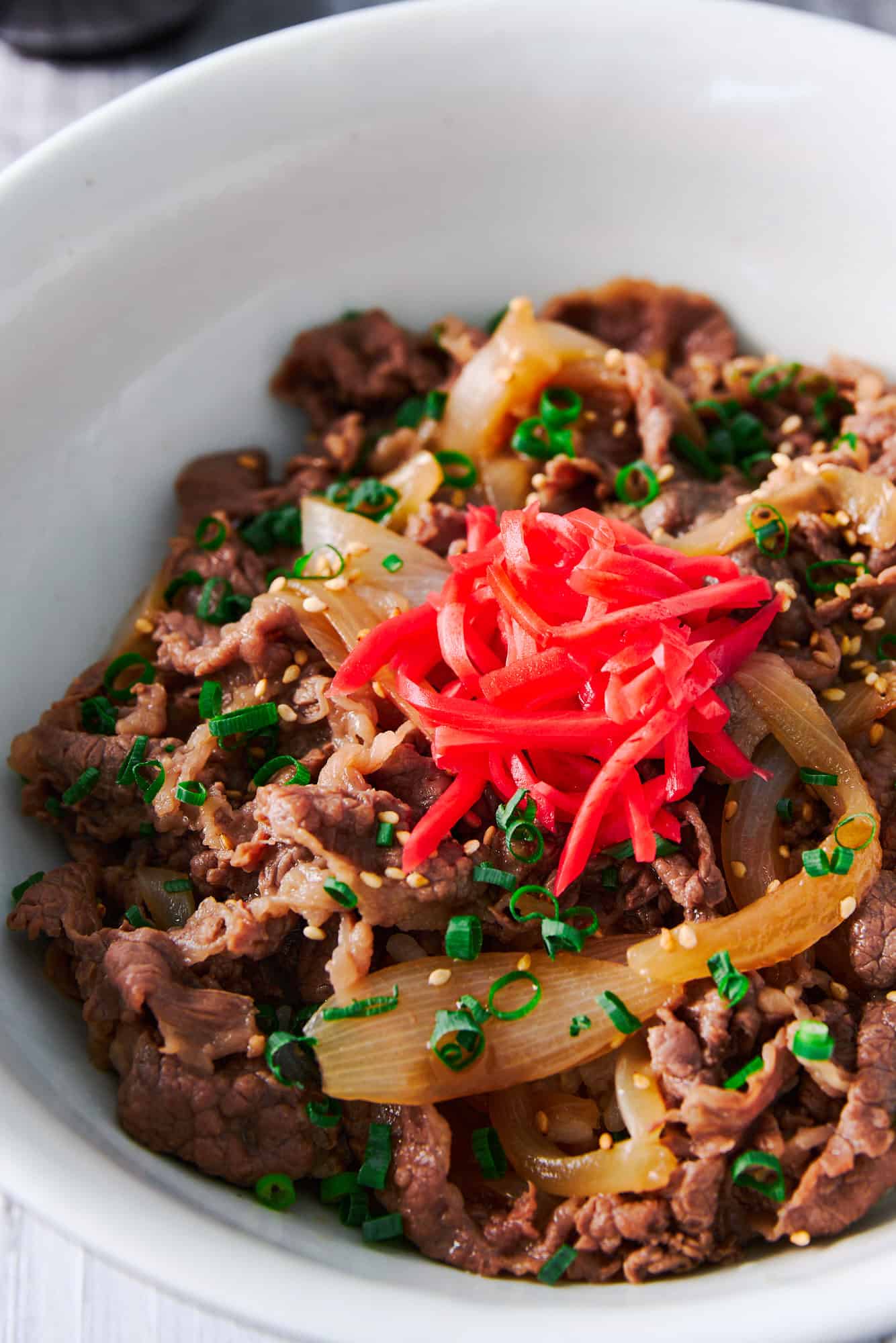 Japanese beef bowl recipe or gyudon with thinly sliced beef simmered with onions in a savory broth, topped with sesame seeds, scallions and red pickled ginger over a bowl of rice.