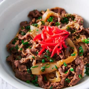 Japanese beef bowl recipe or gyudon with thinly sliced beef simmered with onions in a savory broth, topped with sesame seeds, scallions and red pickled ginger over a bowl of rice.