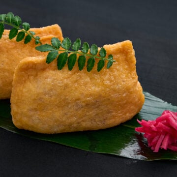 Juicy, umami-rich tofu skins wrapped around gingery vinegared rice, styled as traditional inari sushi with a fresh herb garnish and a bite of beni shoga on the side.