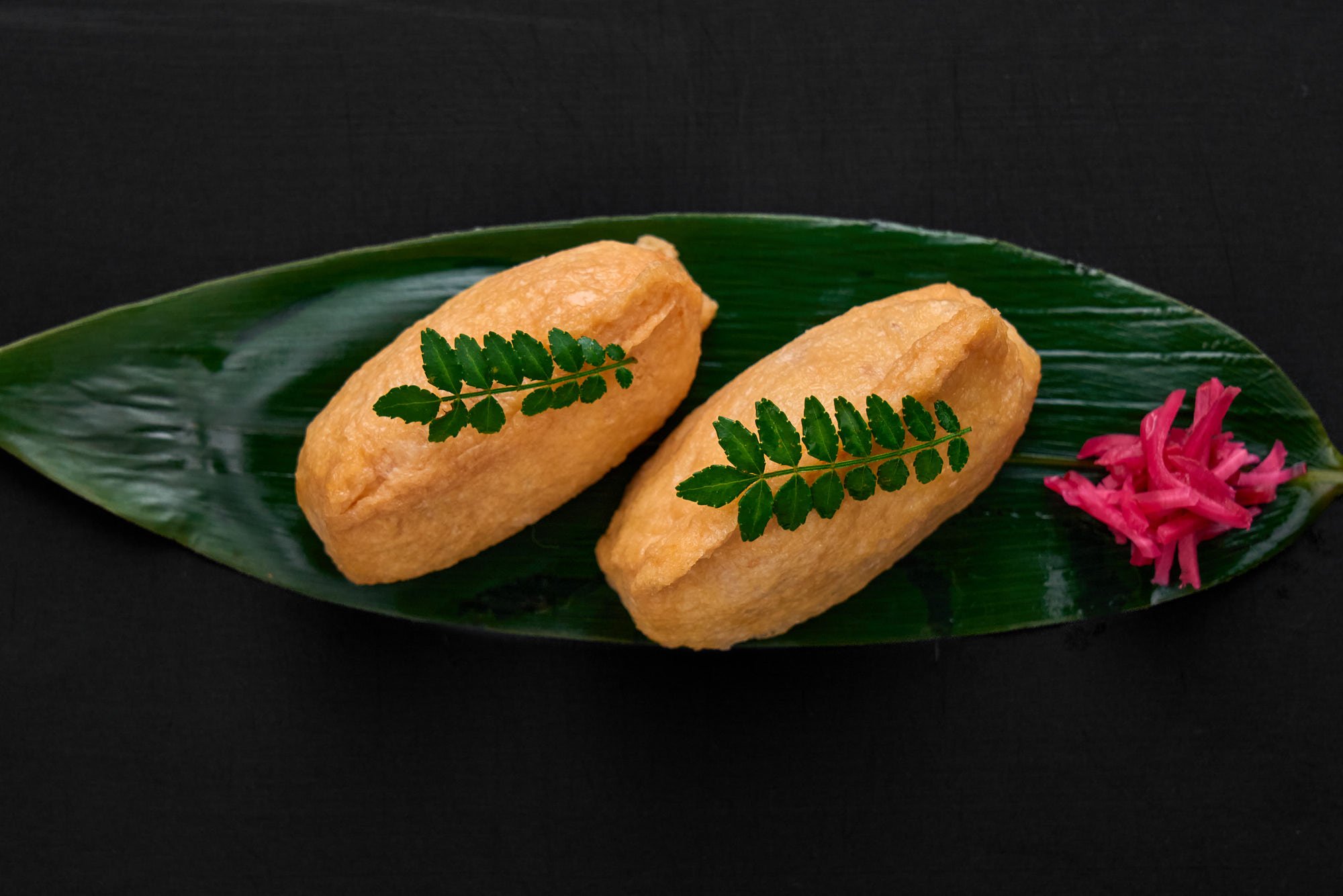 Two plump inari sushi pieces filled with sesame-studded sushi rice, plated simply on a bamboo leaf to highlight the sweet-savory tofu and bright ginger accents.