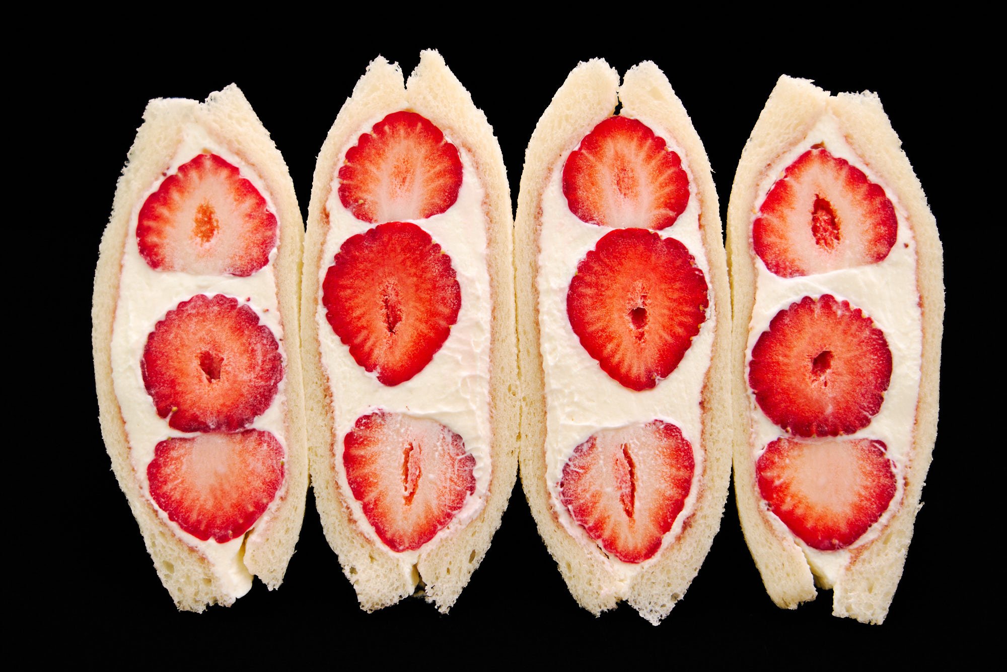 Four beautifully arranged halves of strawberry sandwiches (ichigo sando), showcasing ruby-red berries enveloped in airy whipped cream inside delicately fluffy slices of bread.