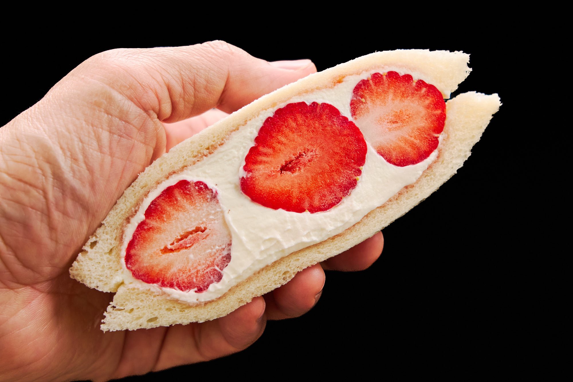 A hand holding an irresistible strawberry sandwich half, displaying vibrant strawberries surrounded by velvety whipped cream and soft, cloud-like bread—ready to enjoy.