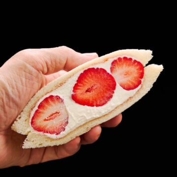 A hand holding an irresistible strawberry sandwich half, displaying vibrant strawberries surrounded by velvety whipped cream and soft, cloud-like bread—ready to enjoy.