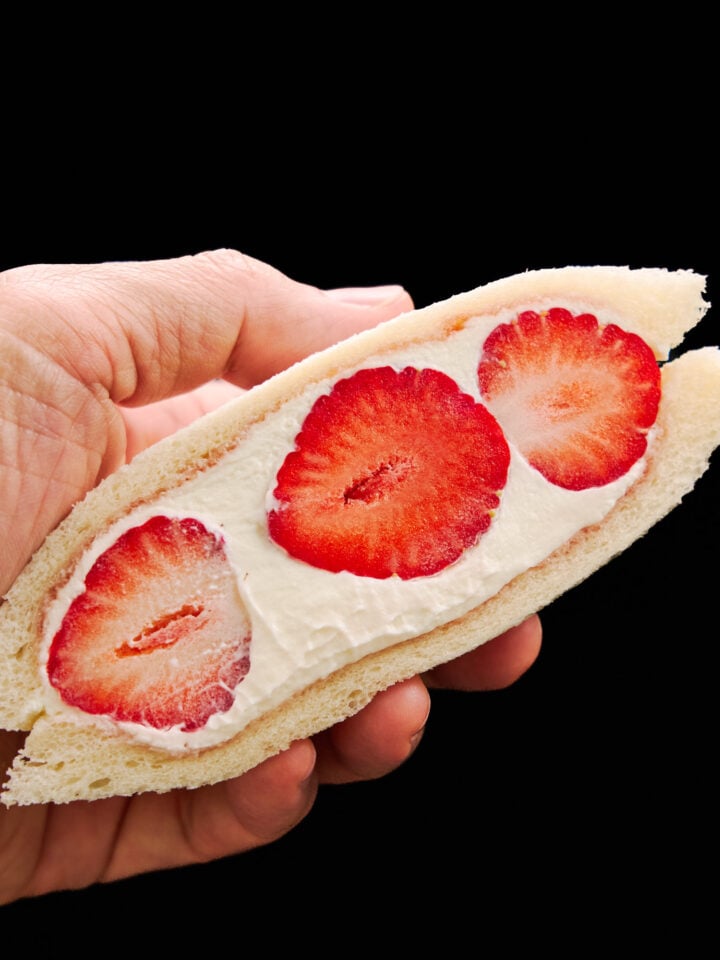 A hand holding an irresistible strawberry sandwich half, displaying vibrant strawberries surrounded by velvety whipped cream and soft, cloud-like bread—ready to enjoy.