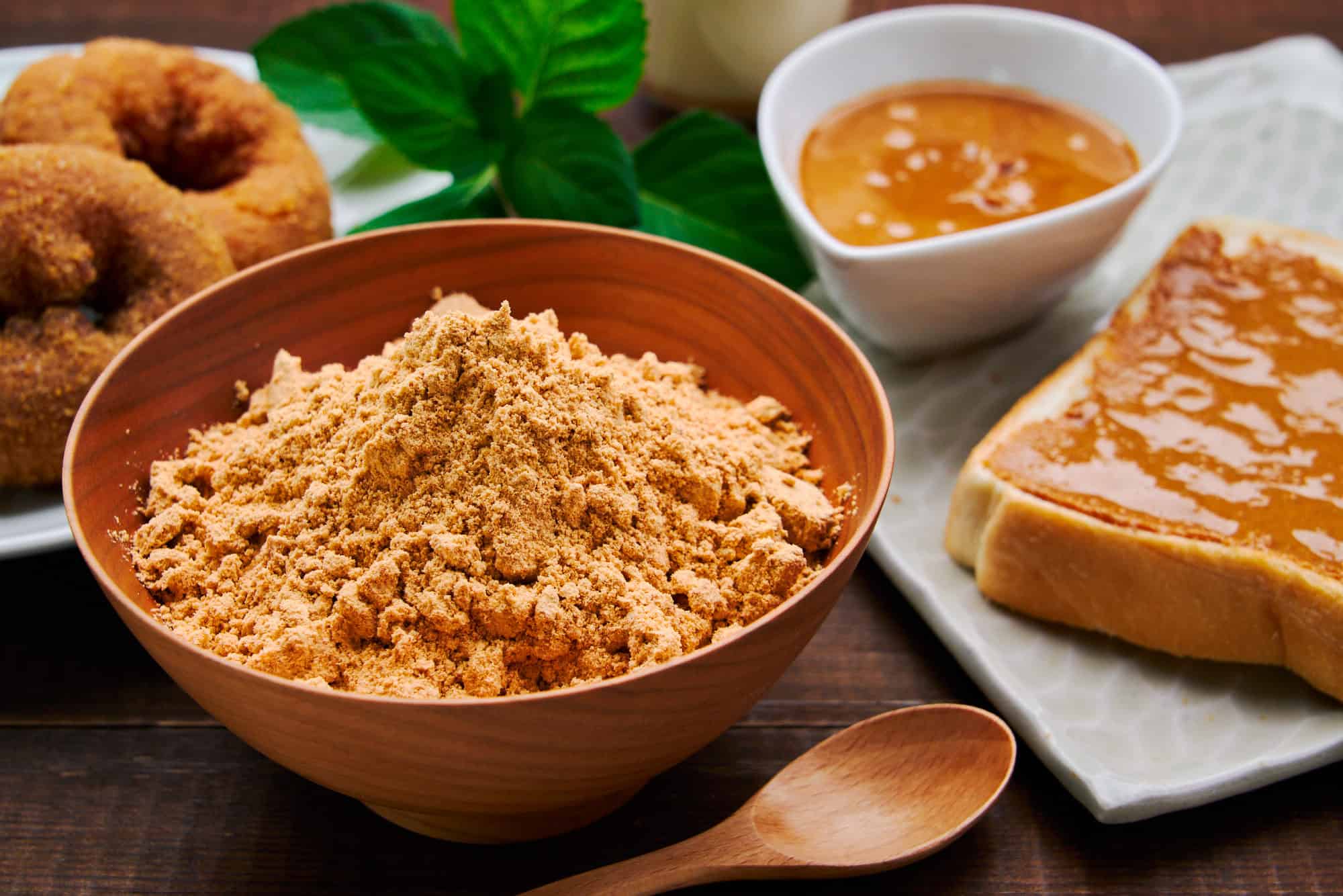 Close-up shot of a wooden bowl filled with homemade kinako powder, its golden hue hinting at its rich, nutty flavor. With kinako doughnuts and kinako butter in the background.
