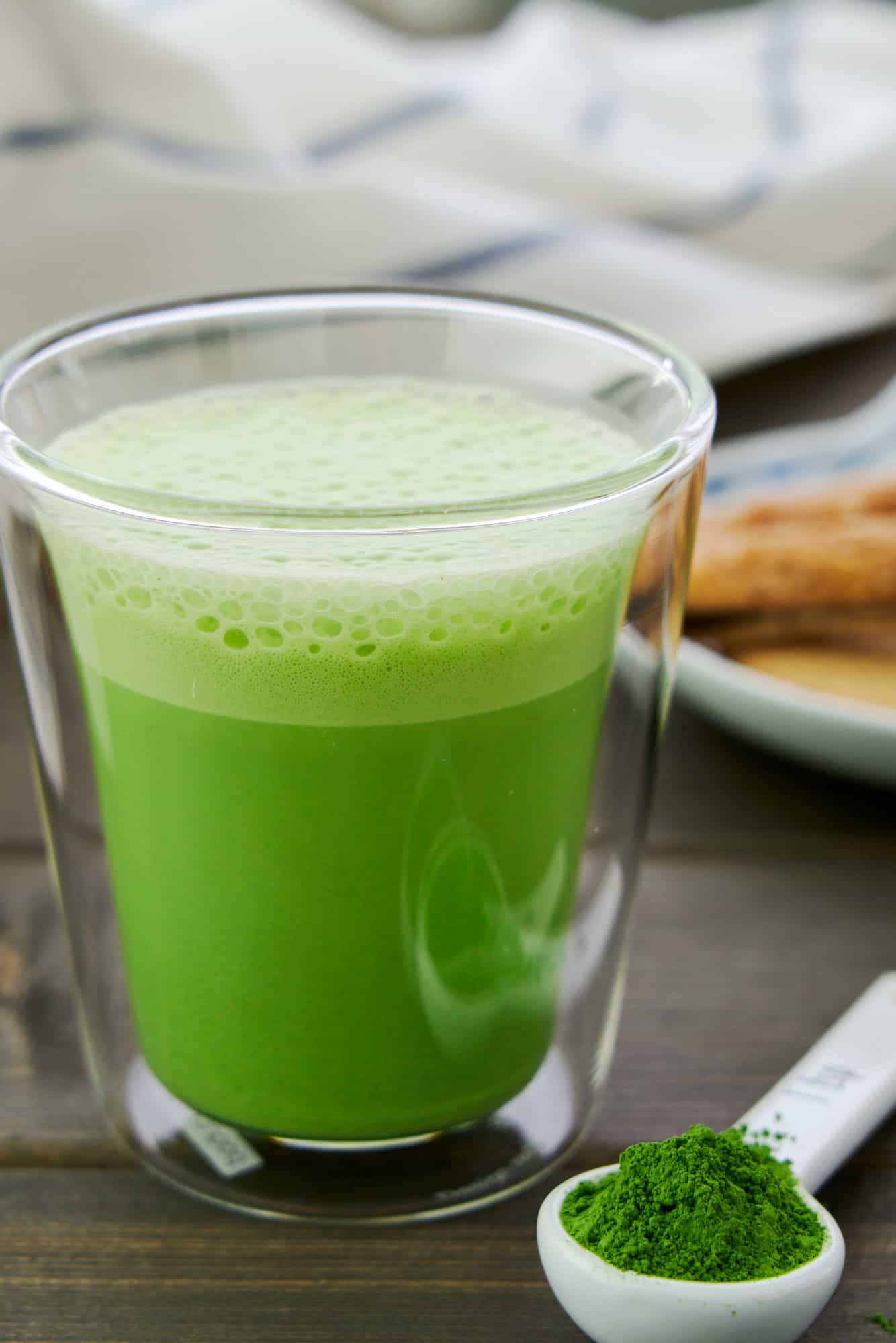 Three ingredient matcha latte recipe makes this glass mug full of creamy Matcha Latte Hot Chocolate. Shown with a spoonful of matcha powder and cookies in the background.