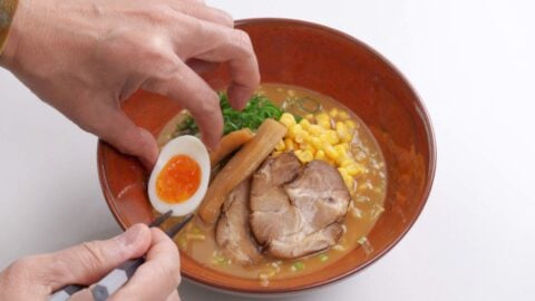 Garnishing miso ramen with various toppings such as egg, chashu, scallions, and corn.