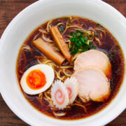 A beautifully plated bowl of shoyu ramen with chicken chashu, soft-boiled egg, menma, narutomaki, and scallions, served alongside rustic wooden chopsticks and spoon.