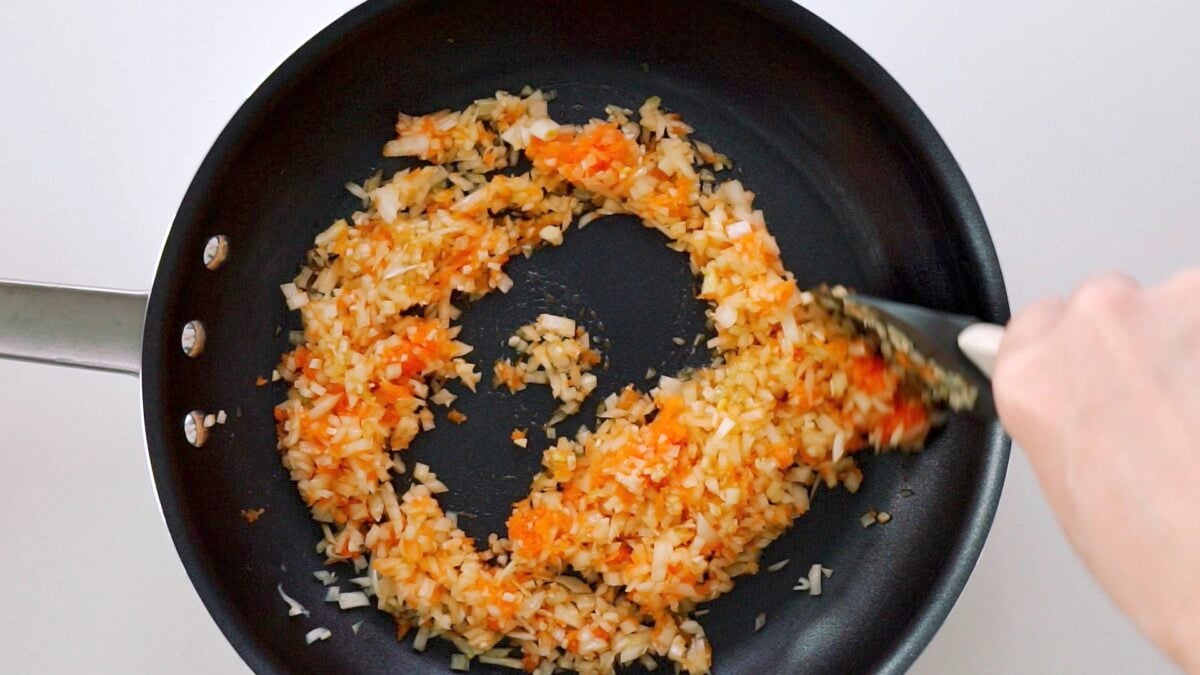 Stirring onions, garlic and grated carrots together in a frying pan.