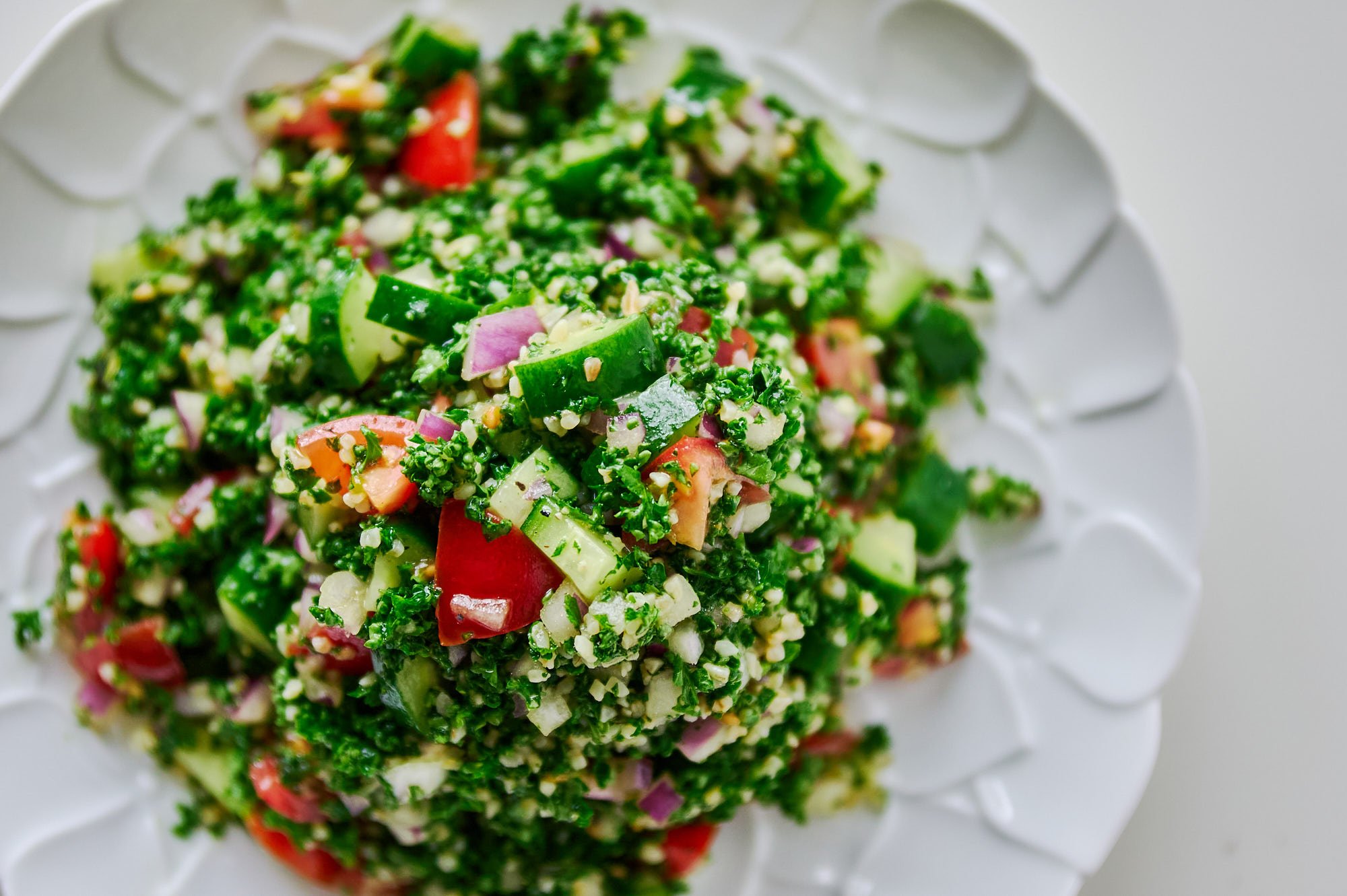 An overhead view of tabbouleh spread across a white platter showcases a lush mosaic of parsley, bulgur, diced tomato, and cucumber ready for serving.