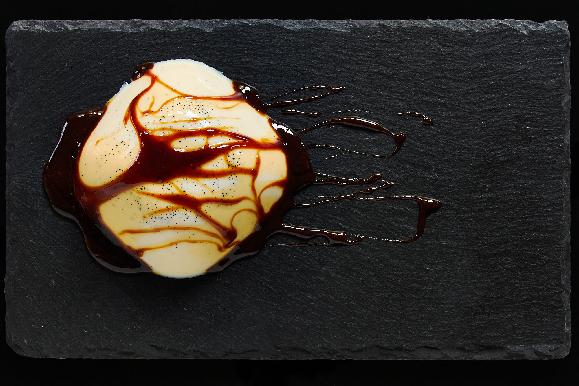 A silky dome of vanilla-specked vegan panna cotta draped with glossy black sugar syrup, artfully served on a slate plate.