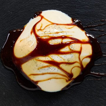 A silky dome of vanilla-specked vegan panna cotta draped with glossy black sugar syrup, artfully served on a slate plate.