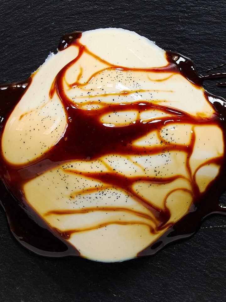 A silky dome of vanilla-specked vegan panna cotta draped with glossy black sugar syrup, artfully served on a slate plate.