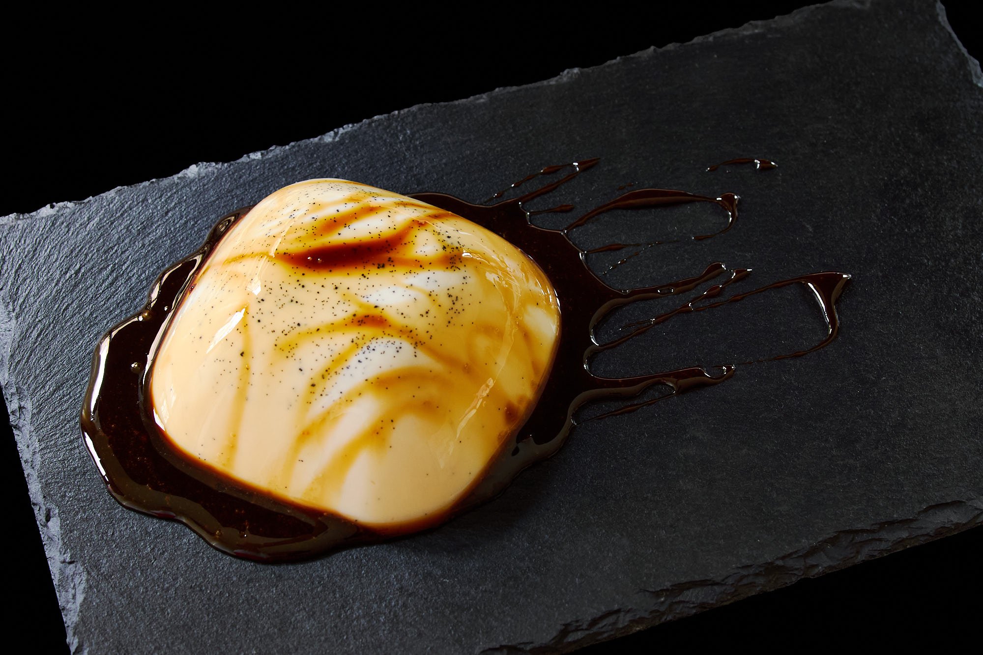 Melt in your mouth plant-based panna cotta glistens with syrupy black sugar sauce, its smooth texture highlighted against the dark slate.