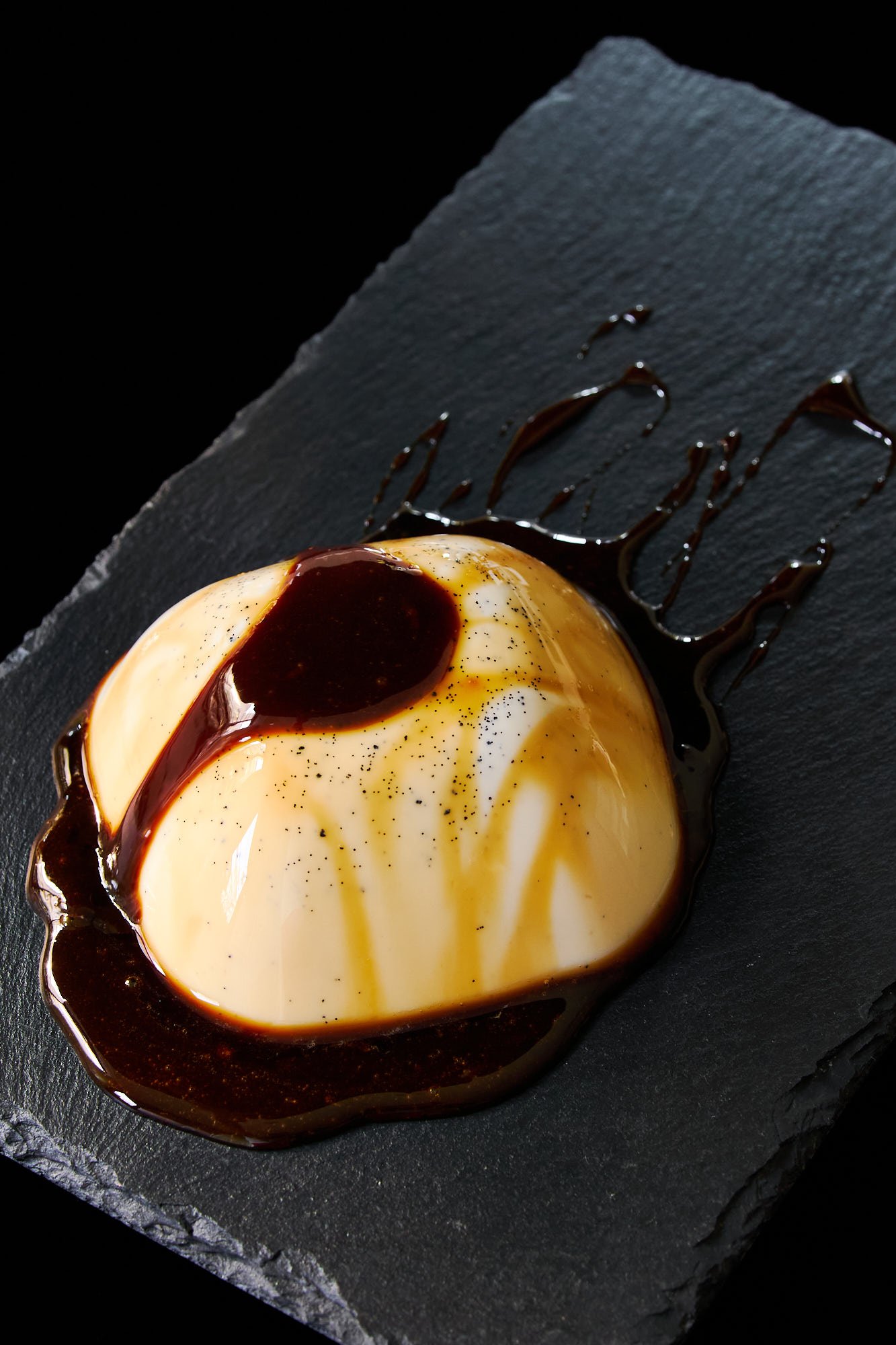 A golden-tinted vegan panna cotta pools in rich black sugar syrup, with an elegant drizzle trailing across the slate for a bold finish.